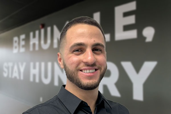 A smiling man in a black shirt stands in front of a wall with the motivational inscription be humble, stay hungry.